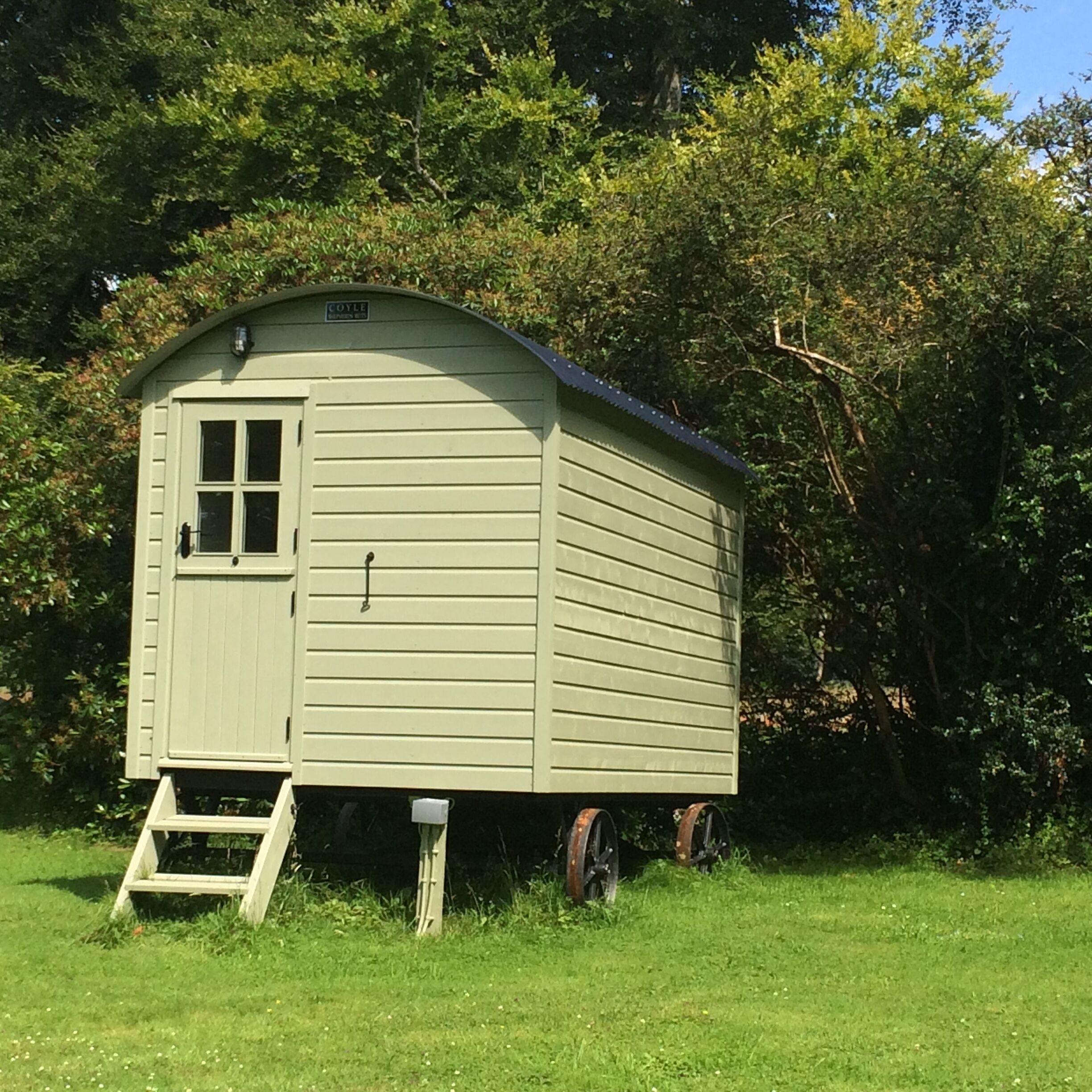 A small, single-story wooden structure with a door and windows, surrounded by grass and trees.