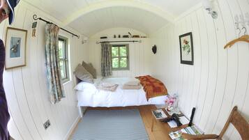 A cozy room with a bed, a small table with a chair, a window with curtains, and a wall-mounted light fixture.