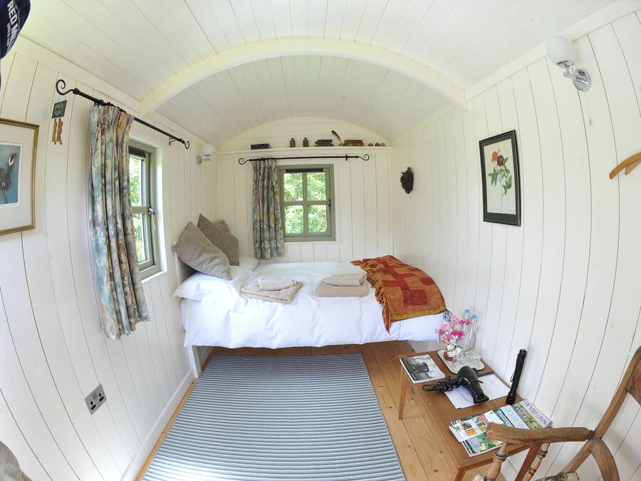 A cozy room with a bed, a small table with a chair, a window with curtains, and a wall-mounted light fixture.