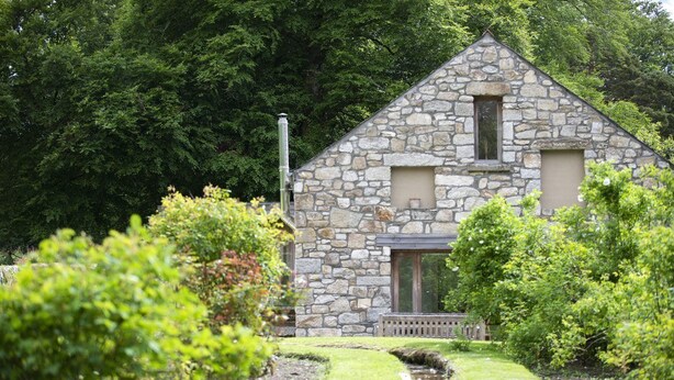A stone building with a triangular roof, surrounded by lush greenery and a small stream. - Blackstairs Shepherds Huts (Knocksquire)