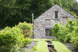 A stone building with a triangular roof, surrounded by lush greenery and a small stream.