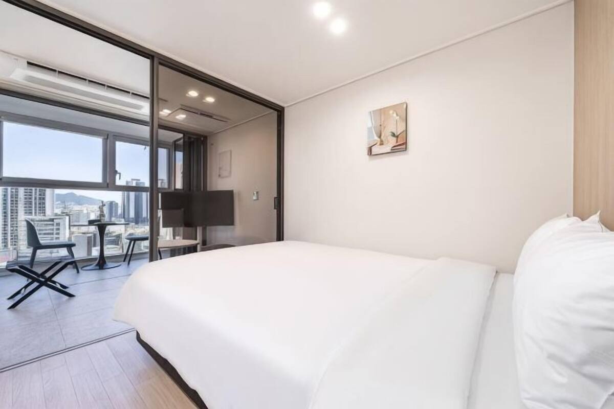 A hotel room with a large bed, a desk, and a chair. There is a TV mounted on the wall and a window with a city view.