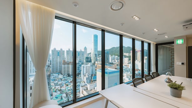 A modern conference room with a large window offering a city view, a white table, and a potted plant.