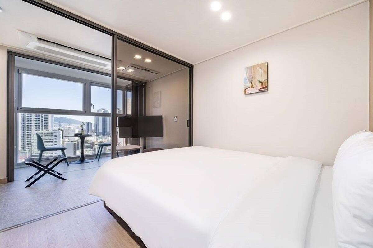 A hotel room with a large bed, a desk, and a chair. There is a window with a view of buildings and a cityscape.
