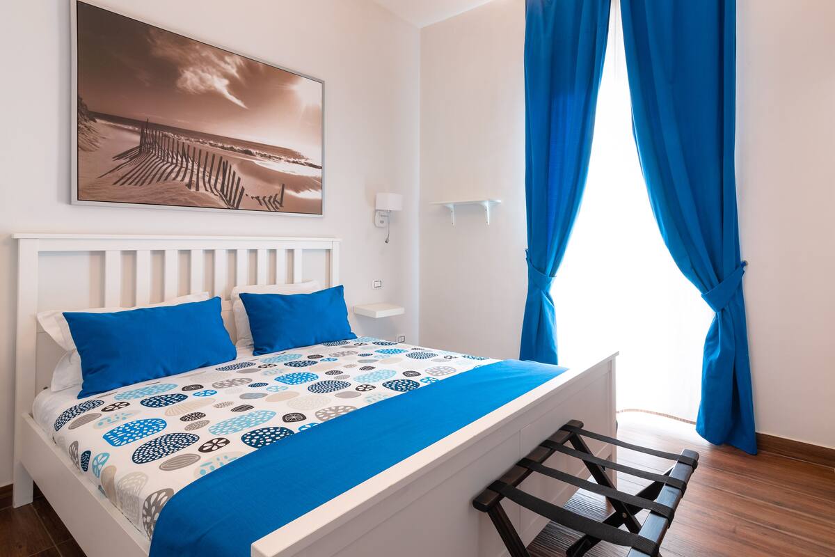 Een slaapkamer met een bed, een hoofdeinde, een wandlamp, een schilderij en blauwe gordijnen.