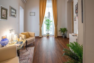 Una sala de estar con un sofá, una mesa de centro con una lámpara, una planta en maceta y vistas de un edificio a través de una ventana con cortinas.