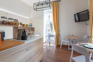 Una cocina moderna con una máquina de café, frascos con aperitivos y una encimera de madera. Hay una zona de comedor con sillas blancas y una mesa, y un televisor montado en la pared.