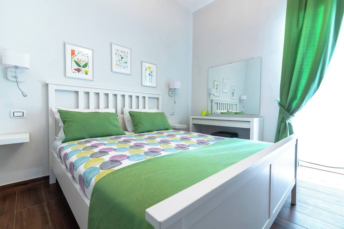 Een slaapkamer met een wit bed, groene kussens, een kleurrijk sprei, een nachtkastje met een lamp en een spiegel.