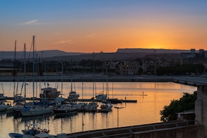 Un puerto deportivo con varias embarcaciones amarradas, un puente al fondo y un atardecer sobre el agua.
