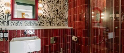 Un baño con pared de azulejos rojos, un lavabo blanco, un inodoro, una ducha y un espejo con marco rojo.