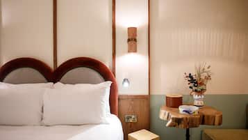 A neatly made bed with white linens, a wooden headboard, and a nightstand with a lamp and a small vase of dried plants.