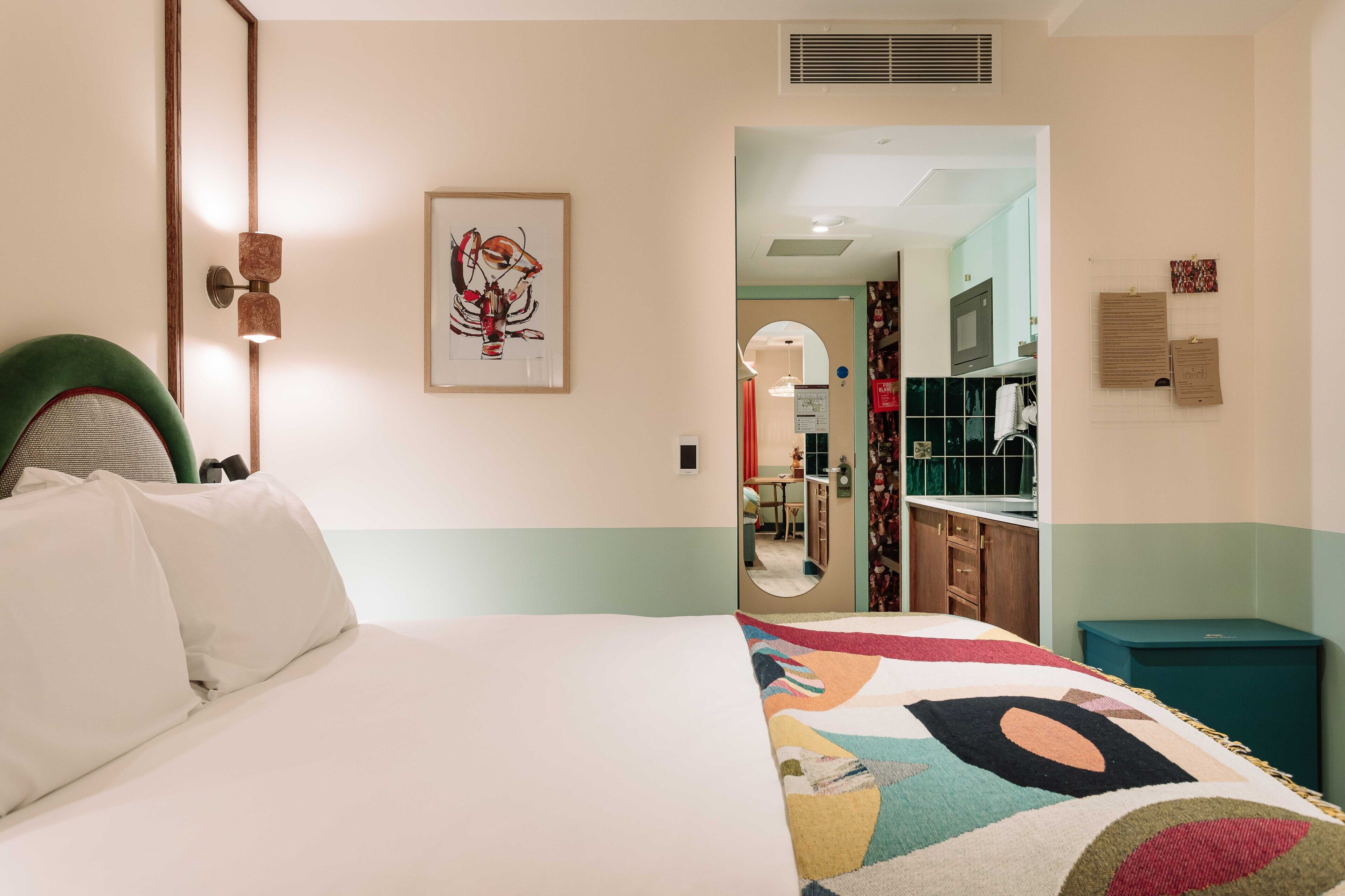 Quarto de hotel com cama, manta colorida, uma obra de arte emoldurada, espelho e vista para a cozinha.