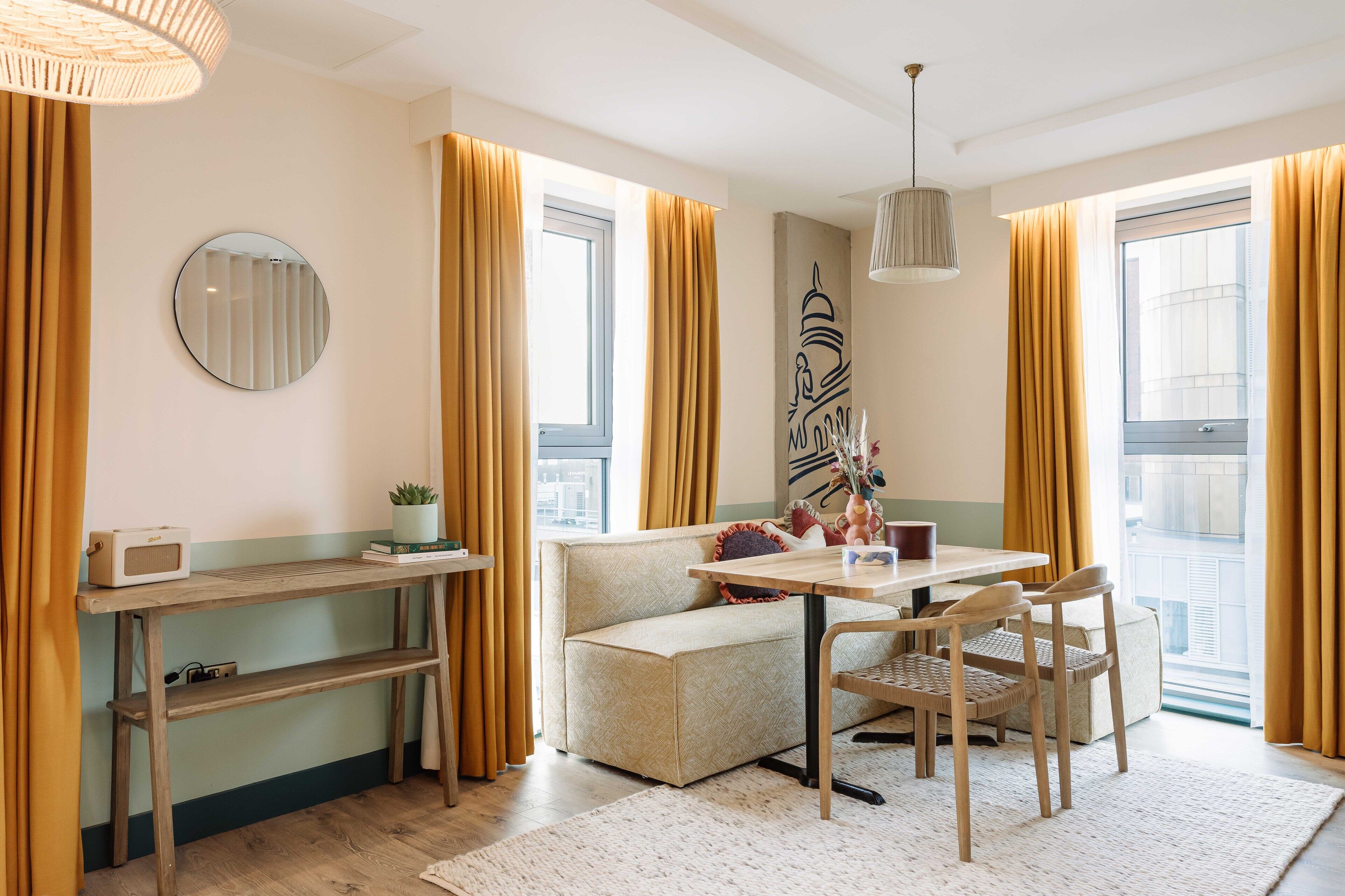 En moderne stue med sofa, spisebord, stole, et rundt spejl og et stort vindue med gardiner.