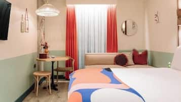 A hotel room with a bed, a small table with a chair, a window with curtains, and a wall-mounted mirror.