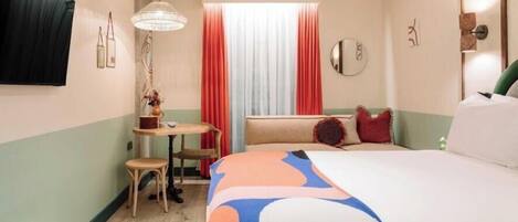 Een hotelkamer met een bed, een klein tafeltje met stoel, een raam met gordijnen en een spiegel aan de muur.