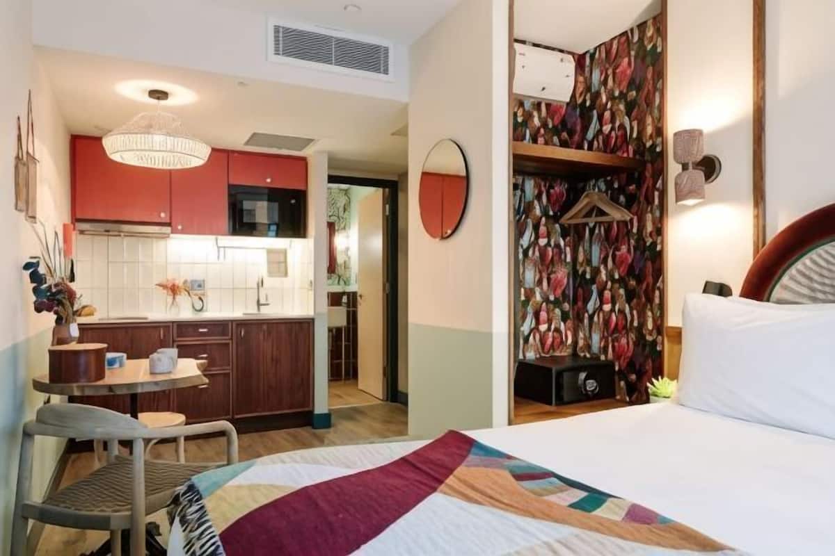 Un dormitorio con cama, escritorio y silla. Cuenta con una zona de cocina con armarios y microondas.