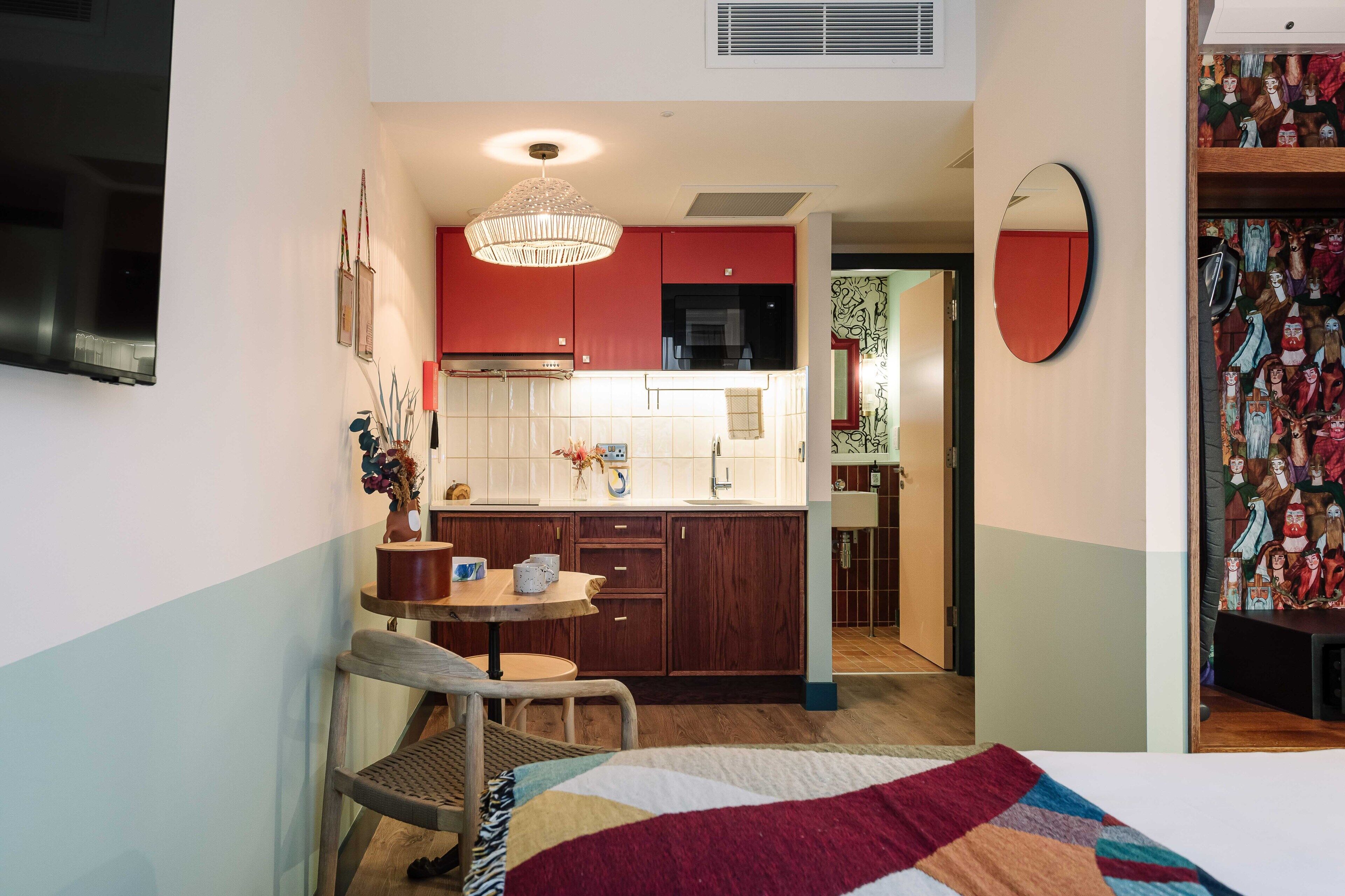 A compact hotel room with a kitchenette, a dining area with a table and chairs, and a bed with a colorful quilt.