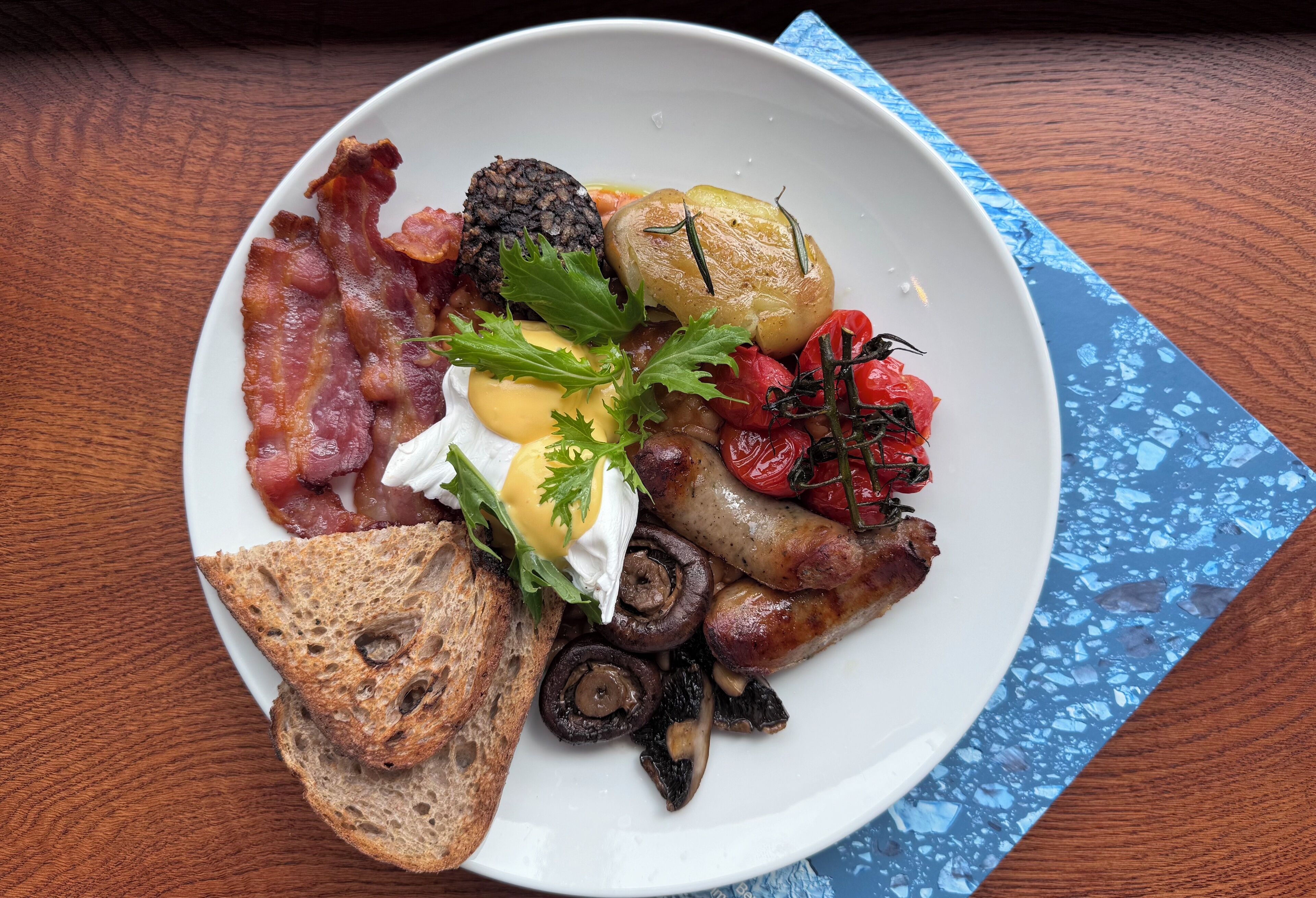 Une assiette remplie de plats pour le petit-déjeuner, comprenant du bacon, des œufs, des champignons, des tomates et des toasts, posée sur une table en bois.