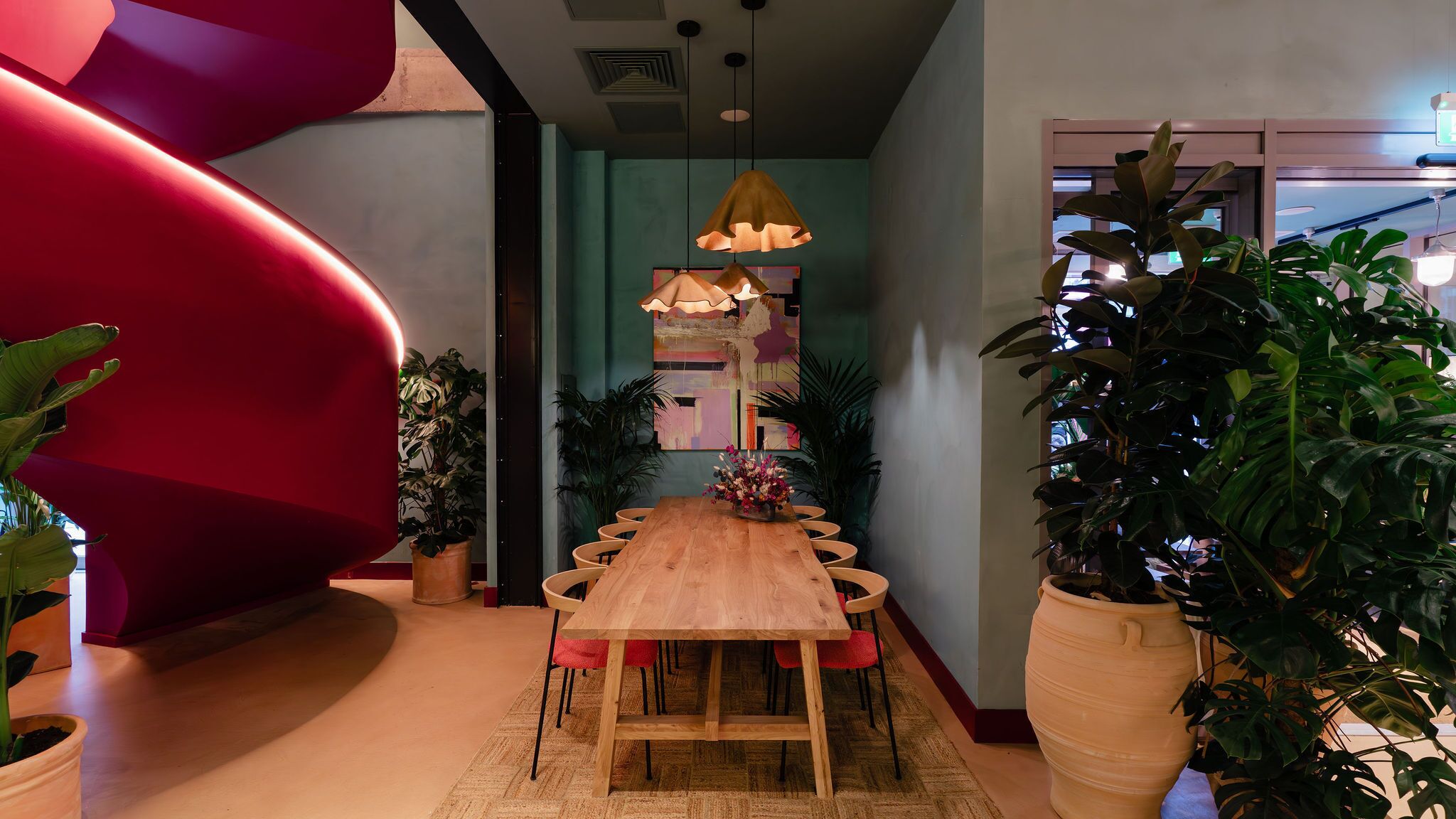 Uma sala de jantar moderna com mesa e cadeiras de madeira, rodeada por plantas em vasos e uma grande escultura vermelha.