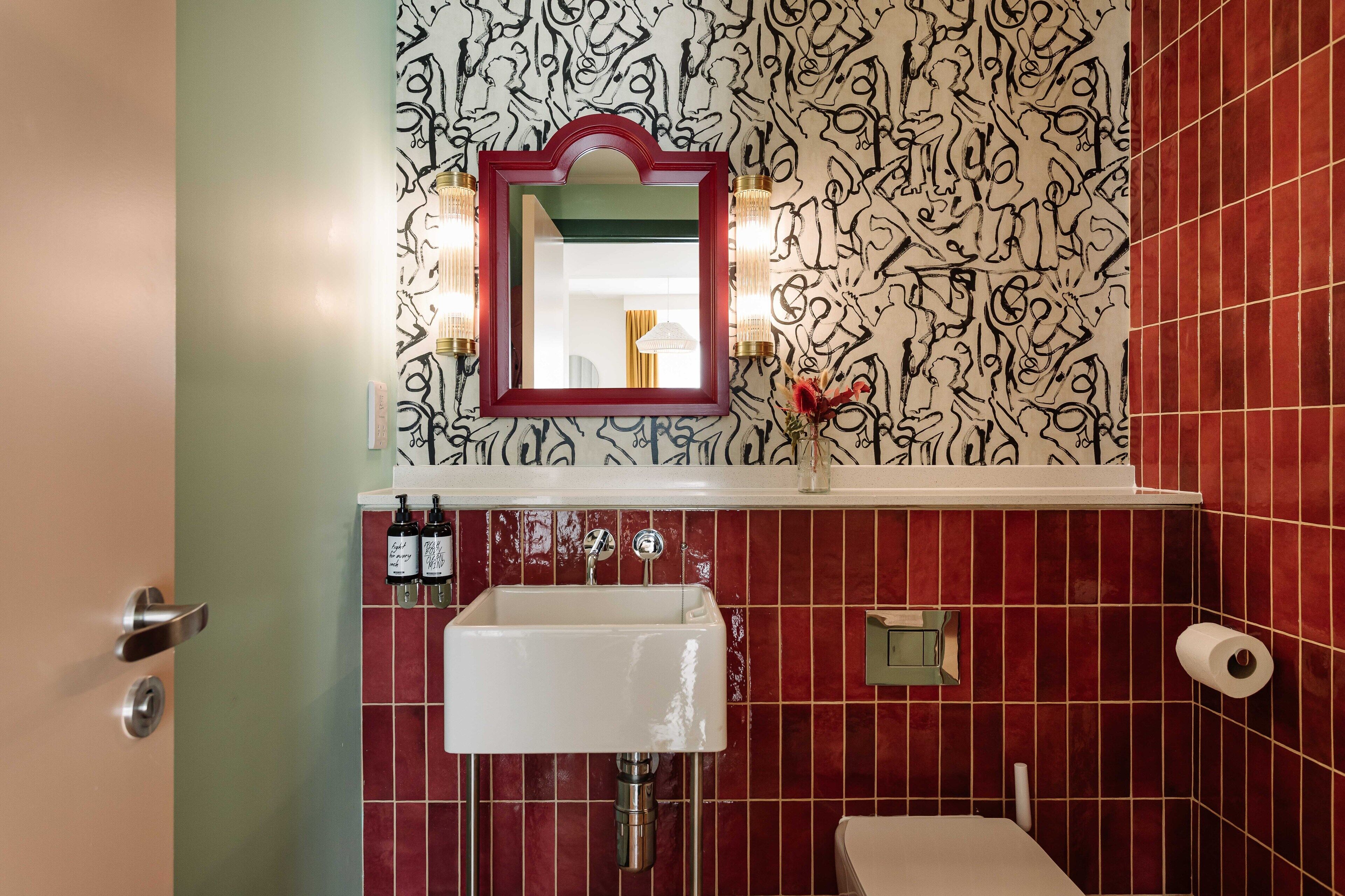 Une salle de bain avec un mur recouvert de carreaux rouges, un lavabo blanc, un miroir à cadre rouge et un petit vase contenant une fleur rouge.