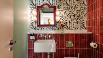 Un baño con pared de azulejos rojos, un lavabo blanco, un espejo con marco rojo y un pequeño florero con una flor roja.