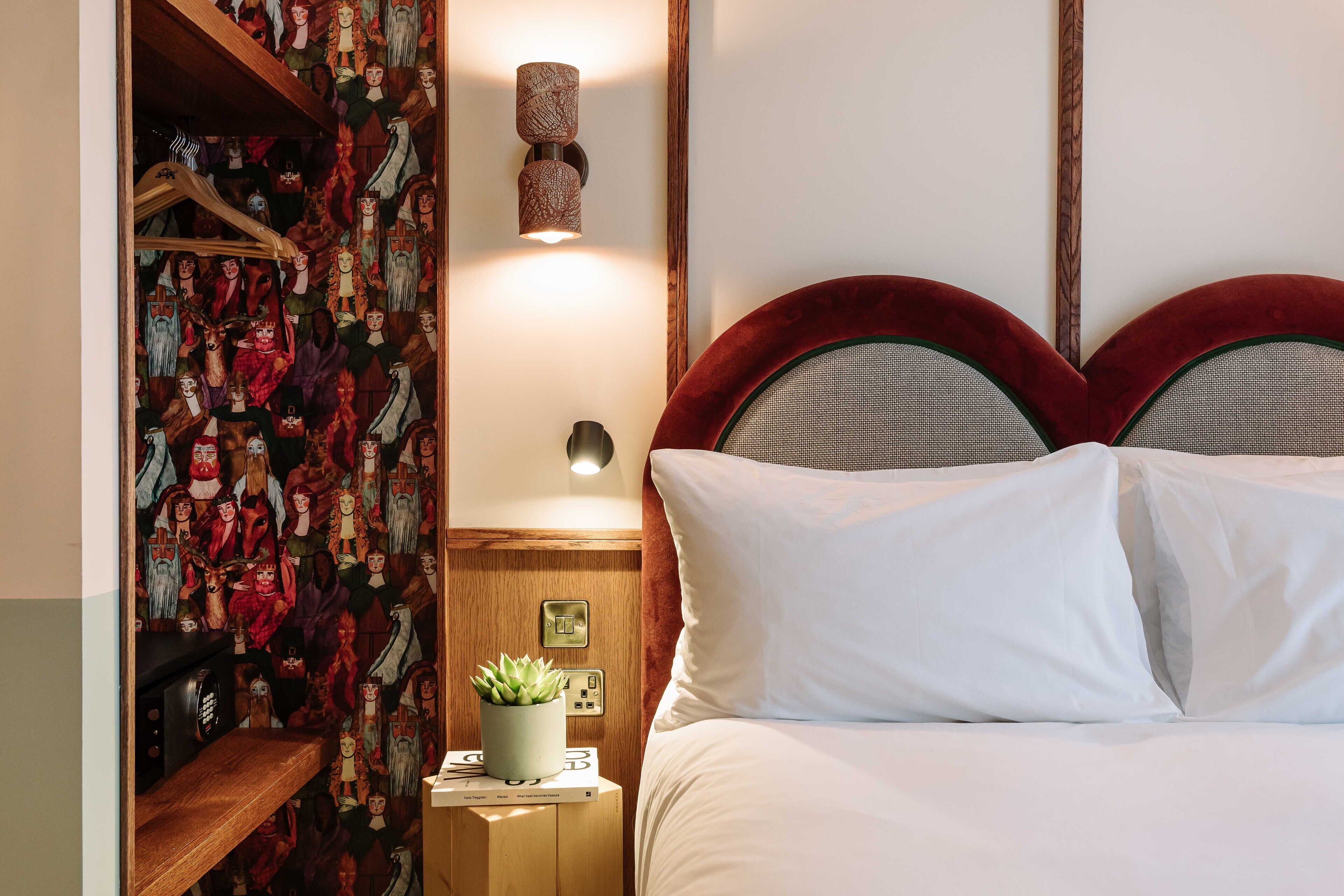 Una habitación de hotel con una cama, una mesita de noche con una lámpara, una pared con papel tapiz estampado y un estante de madera con varios objetos.