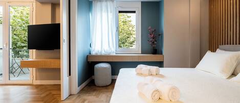 A modern bedroom with a large bed, a TV mounted on the wall, a window with curtains, and a wooden bench by the window.