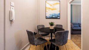 A dining area with a round table, four grey upholstered chairs, a potted plant, and a painting on the wall.