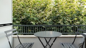 A balcony with a round table and two chairs, surrounded by a metal railing, with a view of dense green foliage in the background.