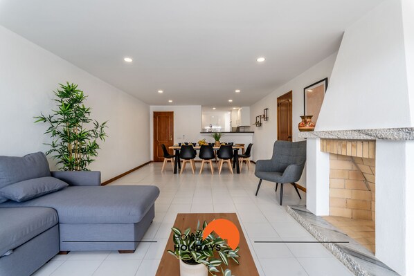 Una sala de estar moderna con un sofá gris, una mesa de centro de madera con plantas, y un comedor con una mesa y sillas de madera.