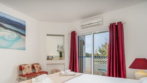 A hotel room with a large painting of a landscape, a bed with white linens, a small table with a lamp, and a balcony with red curtains. - #174 T2 em Zona Tranquila by Home Holidays (Albufeira)