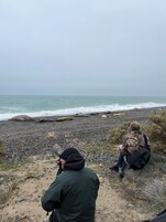 Una playa con costa rocosa, una persona tomando una fotografía y otra persona sentada cerca.