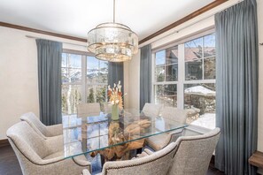 Un espace repas avec une table en verre, des chaises rembourrées, un lustre et une vue dégagée grâce à de grandes fenêtres.