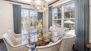 A dining area with a glass table surrounded by six chairs, a chandelier, and a view of a snowy landscape through large windows. - Luxury Mountain Village Retreat | Cassidy Ridge 4BR with Panoramic Views (Mountain Village)