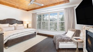 A bedroom with a large bed, a ceiling fan, a fireplace, and a window with a view of trees. - Cassidy Ridge D101 Condo (Mountain Village)