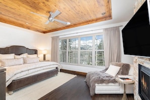 A bedroom with a large bed, a ceiling fan, a fireplace, and a window with a view of trees. - Cassidy Ridge D101 Condo (Mountain Village)