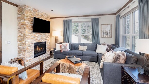 A living room with a stone fireplace, a large flat-screen TV mounted above it, a dark-colored sofa with multiple cushions, and a wooden coffee table. - Cassidy Ridge D101 Condo (Mountain Village)