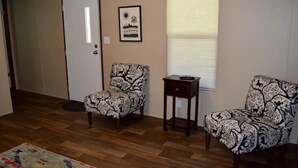 A room with a wooden floor, two patterned armchairs, a small table, a door, and a window with blinds.