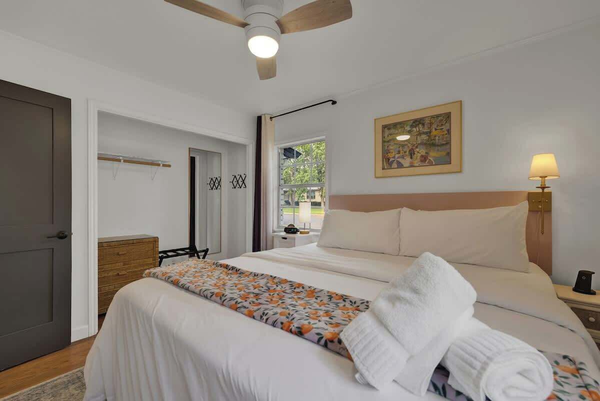 A hotel room with a bed, a bedside table, a lamp, a painting on the wall, and a window with a view of greenery.