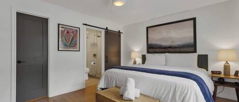 Habitación de hotel con cama, ventilador de techo, un cuadro en la pared, una mesita de noche con lámpara y una puerta que da a otra habitación.