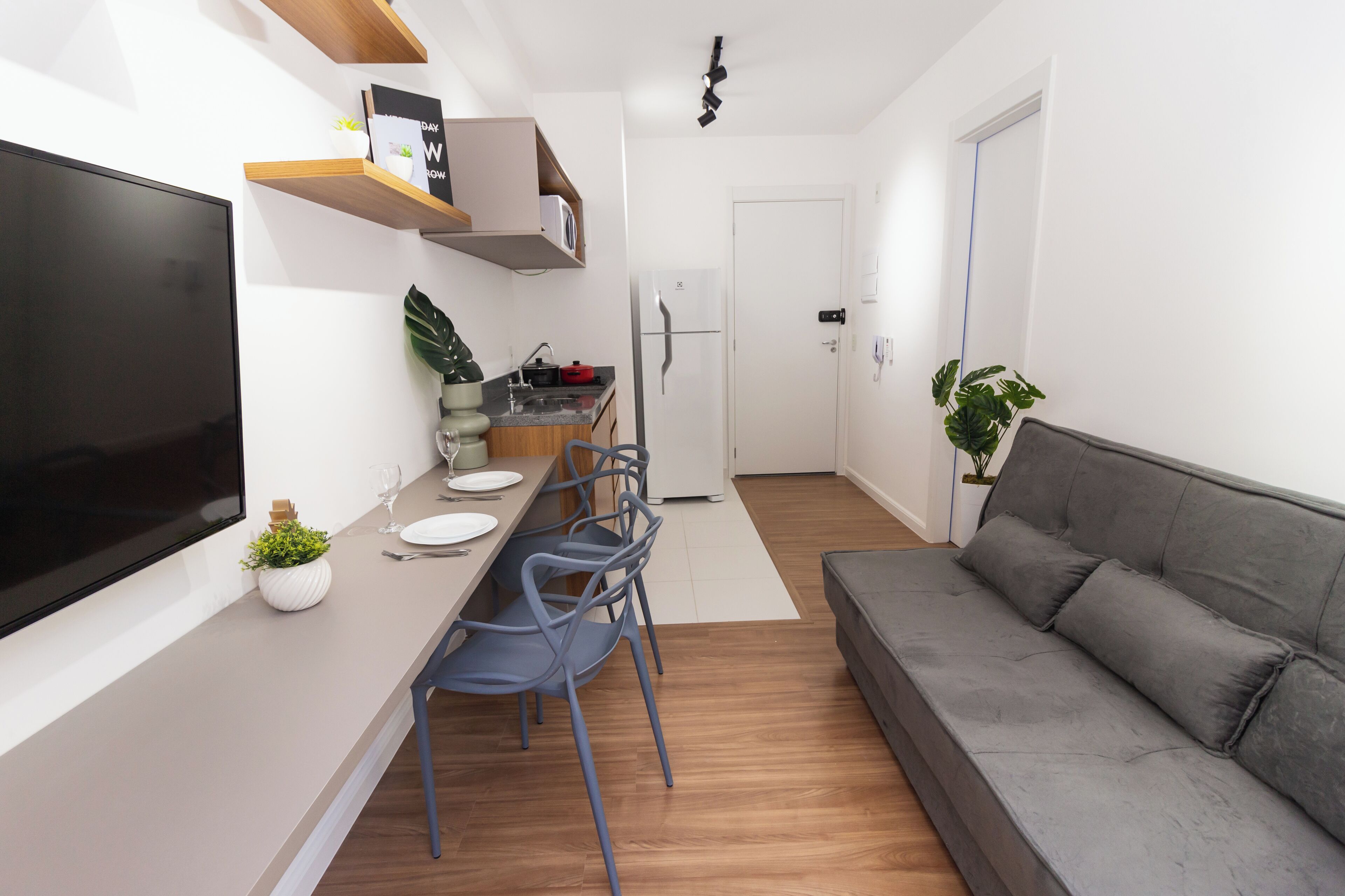 A modern kitchen with a flat-screen TV, wooden shelves, a dining table with chairs, a refrigerator, and a grey sofa.