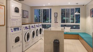 A laundry room with multiple washing machines, a seating area with a bench and cushions, and a trash bin. - 360 For Consolação (São Paulo)