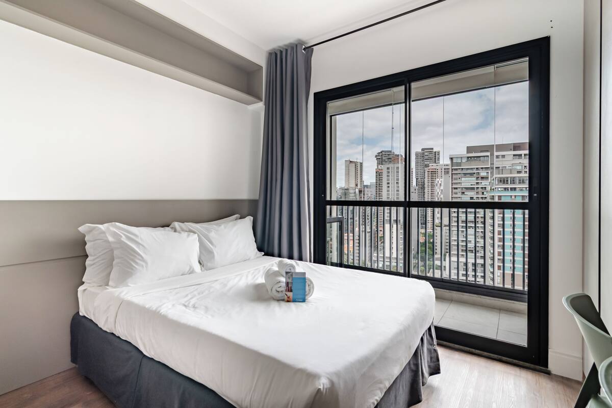 Une chambre d’hôtel moderne dotée d’un grand lit, offrant une vue sur le paysage urbain grâce à une grande fenêtre, et d’un balcon avec une balustrade.