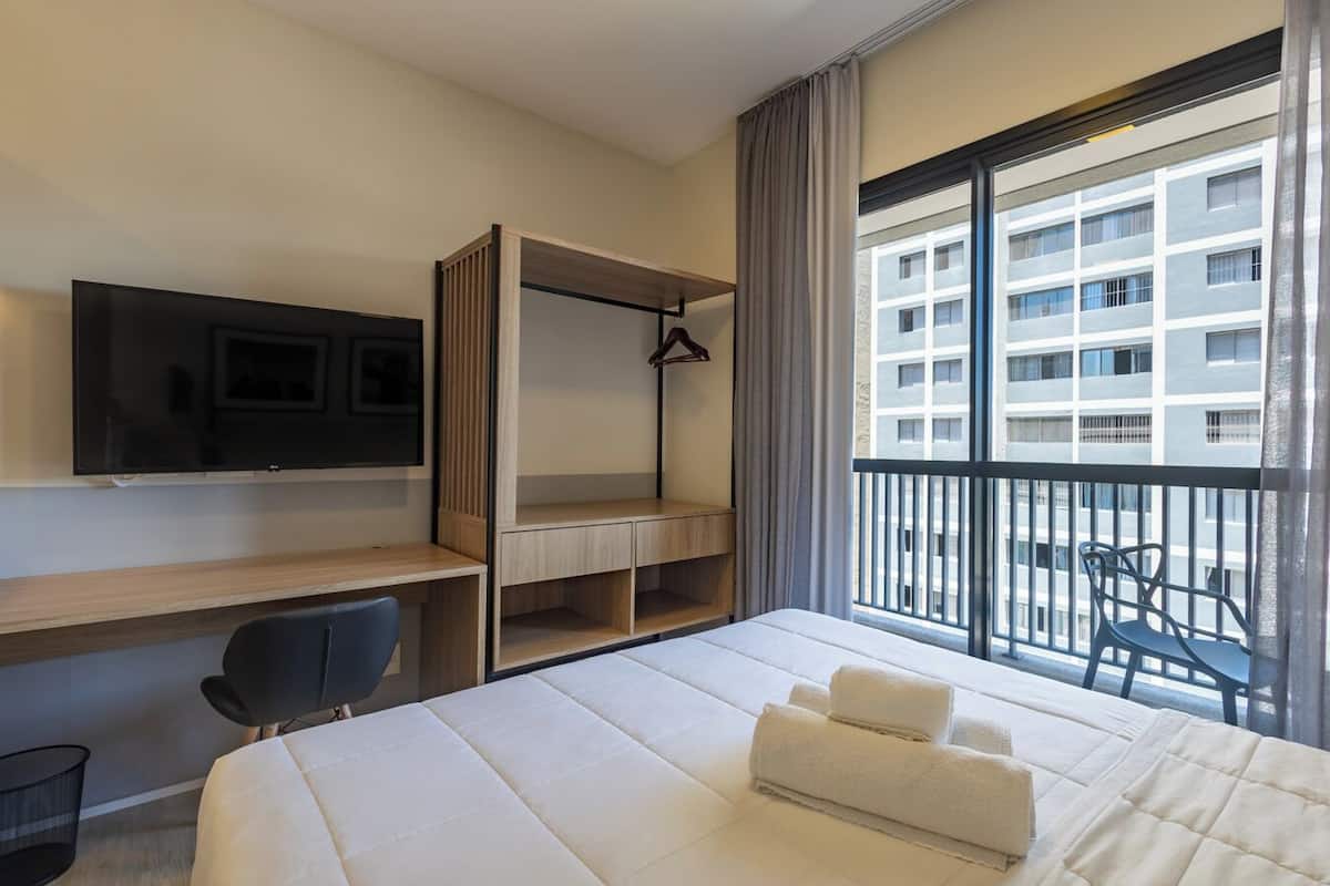 Une chambre d’hôtel comprenant un lit, un bureau, une chaise, une télévision, une armoire et un balcon avec vue sur un immeuble.