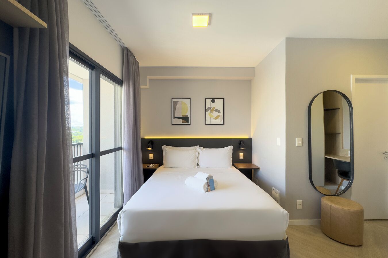 A hotel room with a large bed, a bedside table, a nightstand, a mirror, and a view of the outdoors.