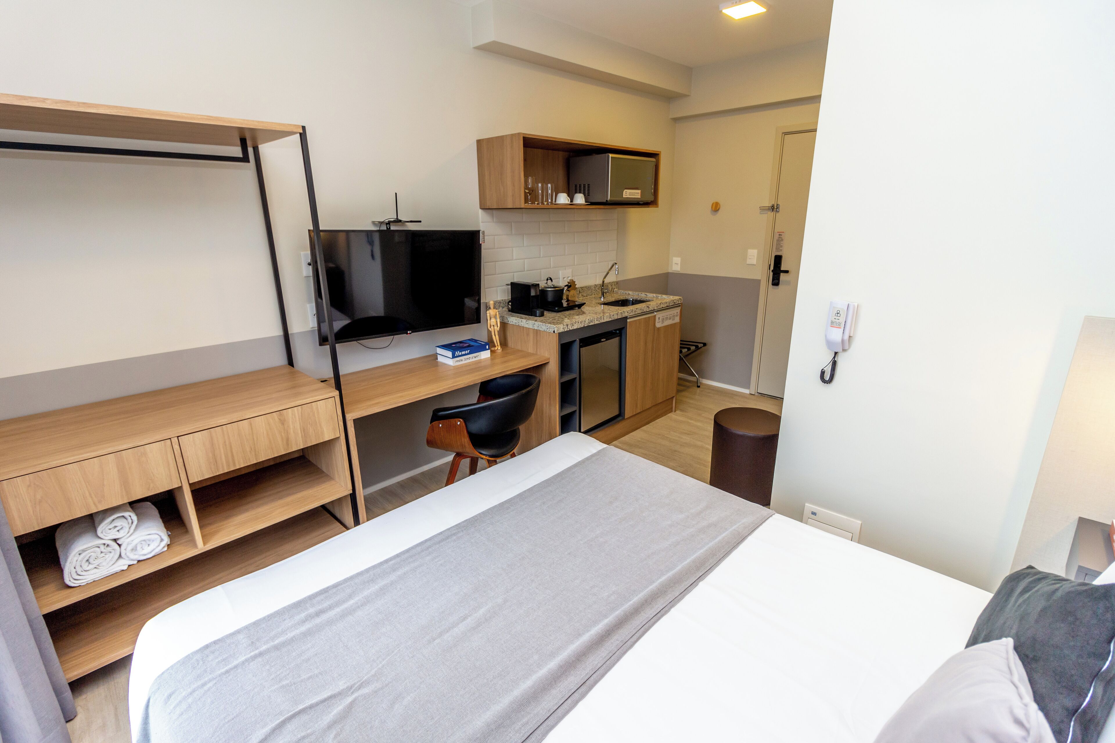 A compact hotel room with a bed, a desk with a chair, a small kitchenette, and a TV.