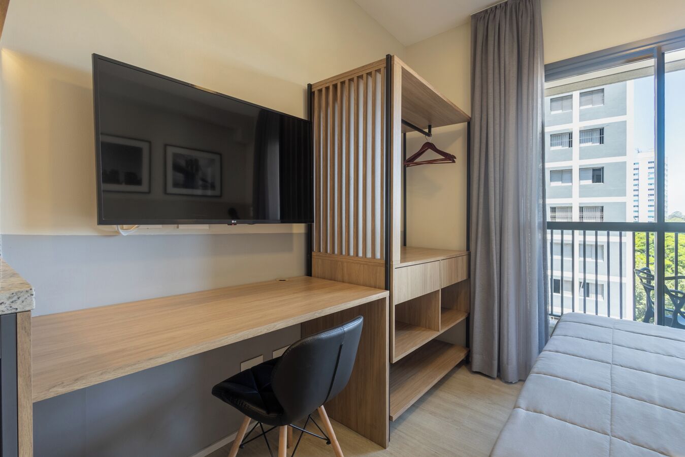 A modern hotel room with a wooden desk, a black chair, a TV mounted on the wall, and a balcony with a view of buildings.