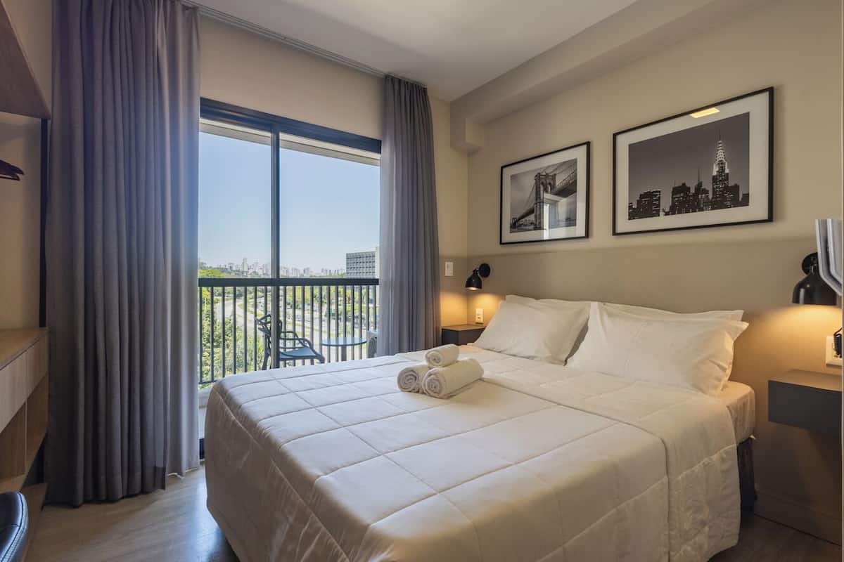 Une chambre d’hôtel avec un grand lit, un balcon avec des rideaux et deux tableaux encadrés au mur.