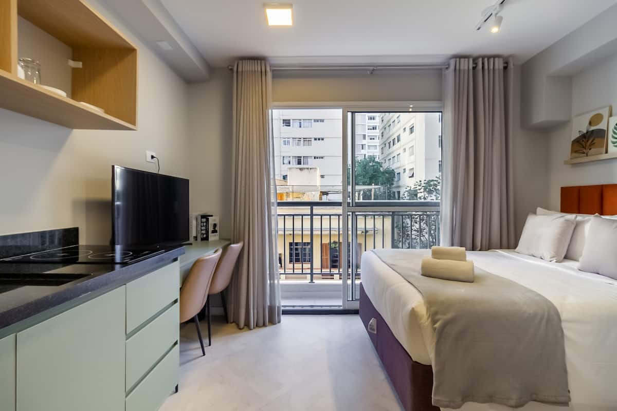 Ein Hotelzimmer mit Bett, Schreibtisch, Fernseher und einem Balkon mit Blick auf Gebäude.