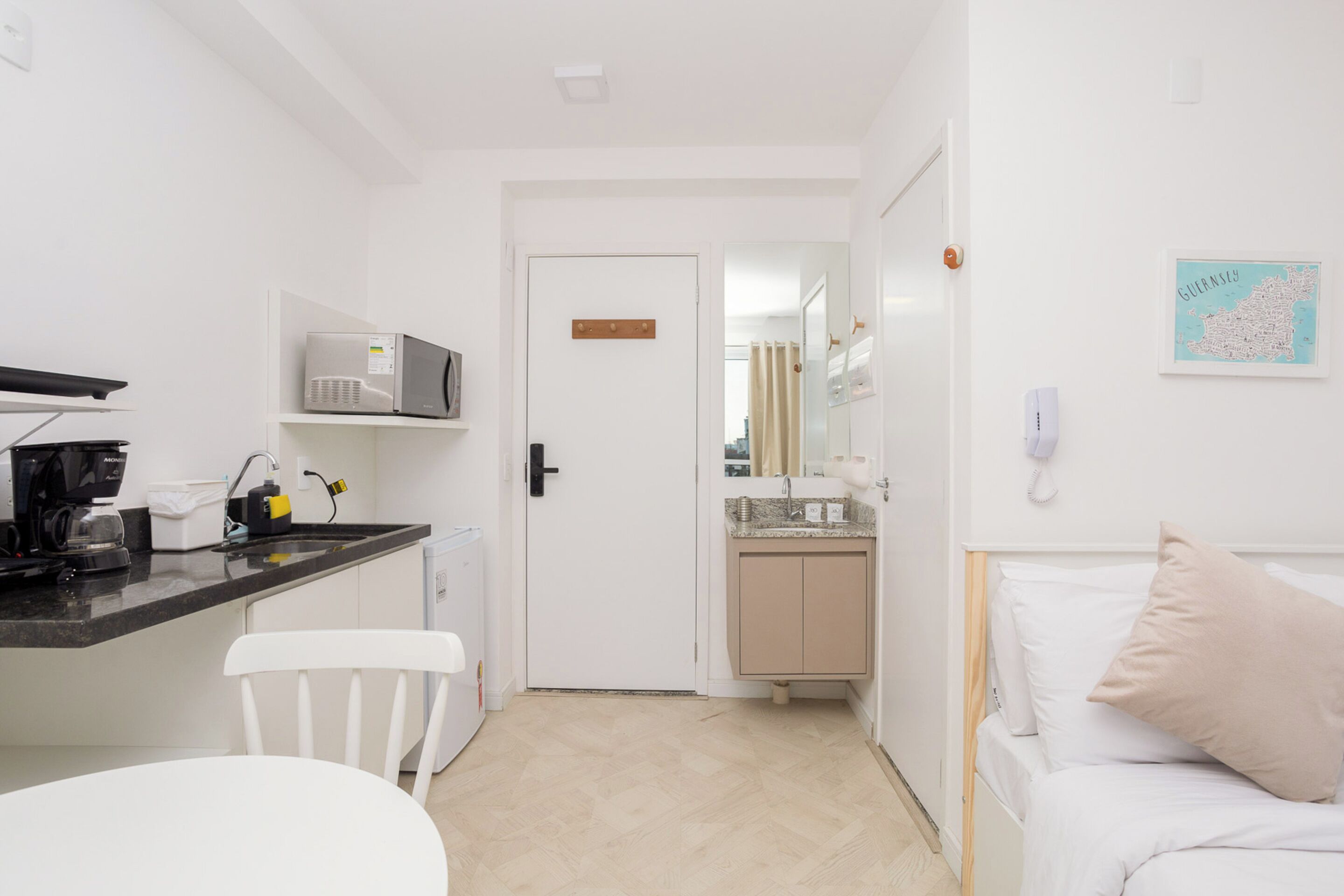 Quarto de hotel pequeno com cozinha compacta, uma cama com um travesseiro, uma cadeira branca e um mapa na parede.